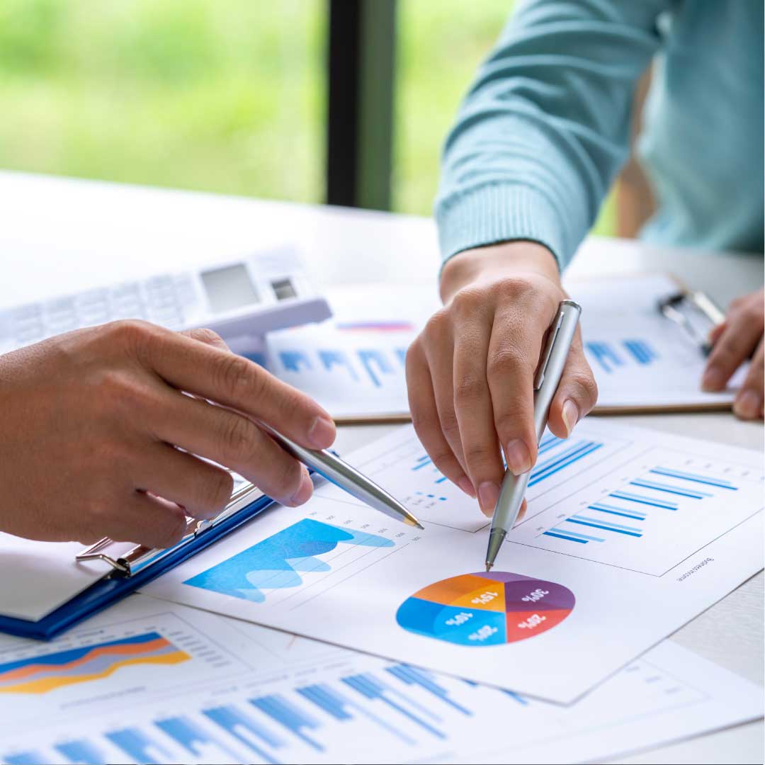 A man and a woman are seated at a desk across from each other, using pens to point at financial data printed on sheets of paper in a well-lit office space.